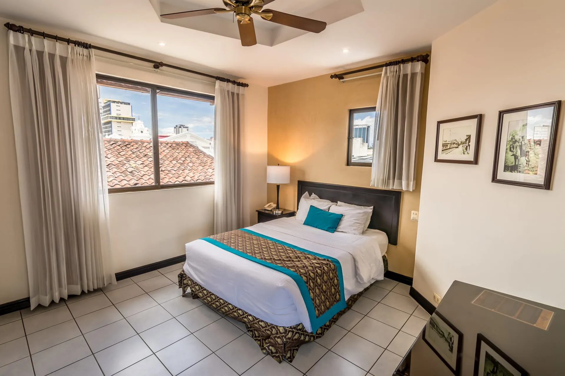 Habitación Doble del Hotel Colonial: cama doble con cabecera oscura, ventilador de techo y ventana con vista a los tejados de teja del centro histórico de San José